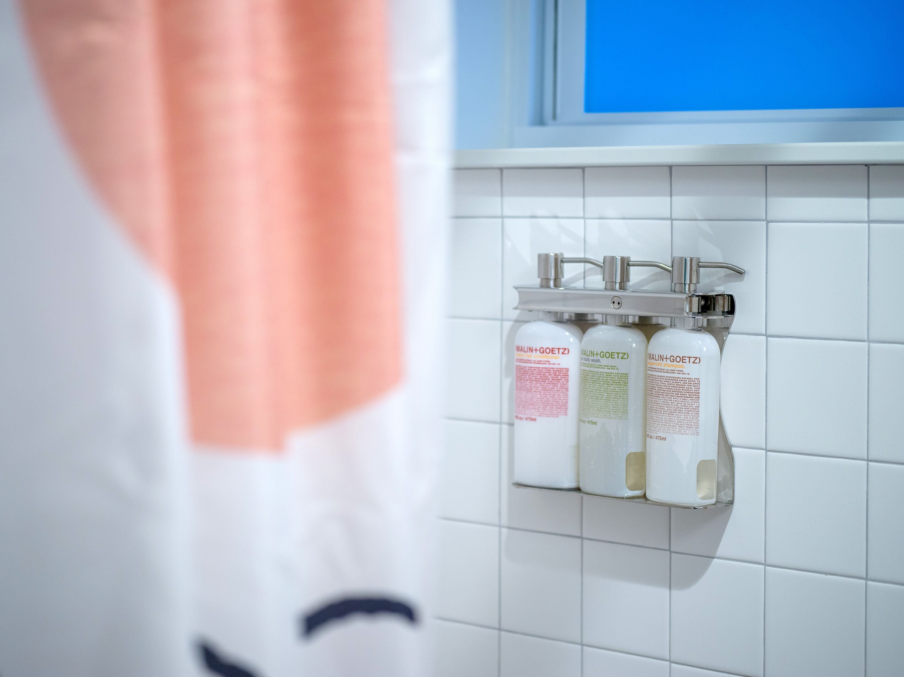 A bathroom with white tiles and a shelf holding three bottles of shampoo.