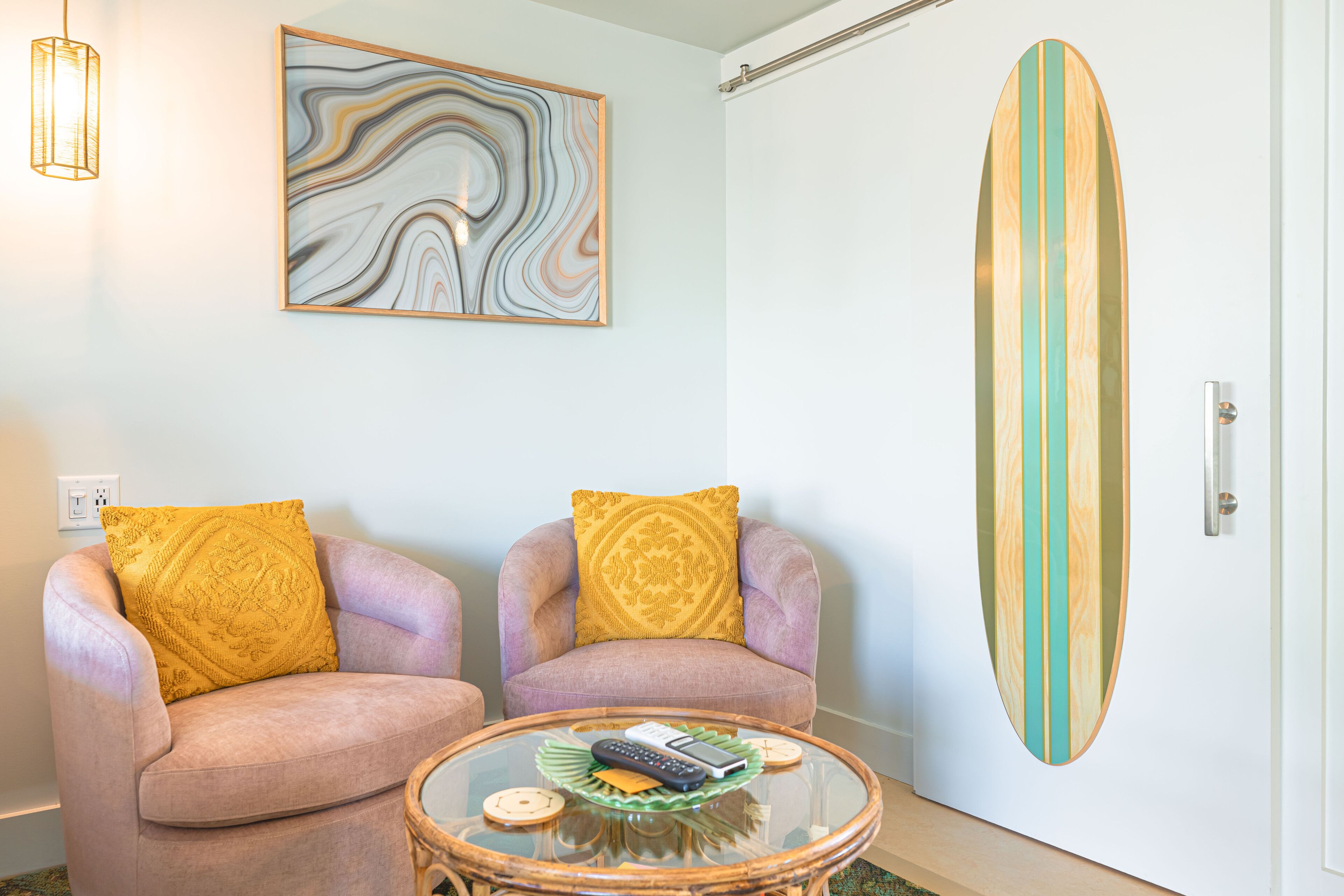 A room with two armchairs, a glass coffee table, a surfboard, and a wall art piece.