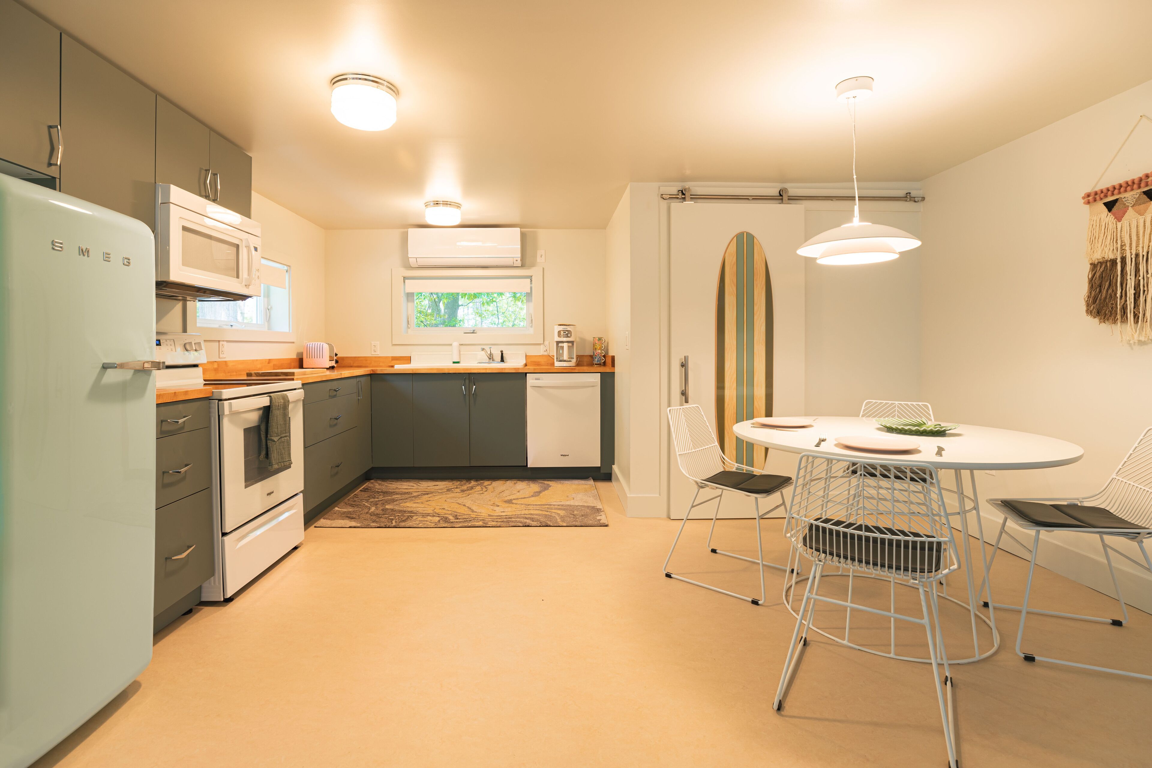 A compact kitchen with modern appliances, a dining area with a round table and chairs, and a surfboard decoration on the wall.