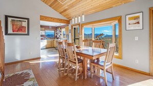 A dining area with a wooden table and chairs, a kitchen in the background, and a large window with a mountain view. - Stargazer Retreat • Hot Tub•Game Room• Creek Walk•DogFriendly• 360° Mtn Views (Nathrop)
