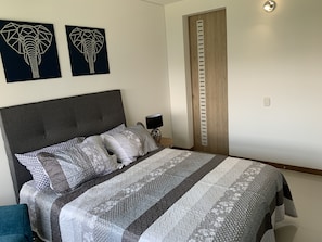 Cama bem arrumada com roupa de cama estampada, cabeceira cinza e duas obras de arte de elefantes penduradas na parede.