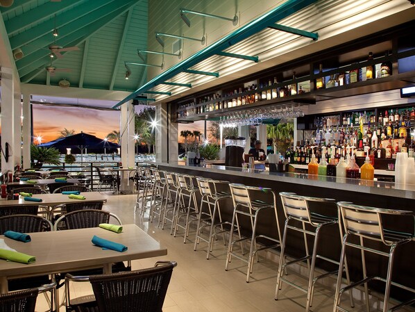 A bar area with a view of the sunset, tables with chairs, and a variety of bottles on shelves.