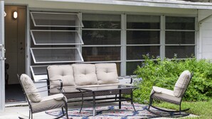Eine Terrasse mit einer Couch und zwei Schaukelstühlen, einem gemusterten Teppich und einem Fenster mit Jalousien.