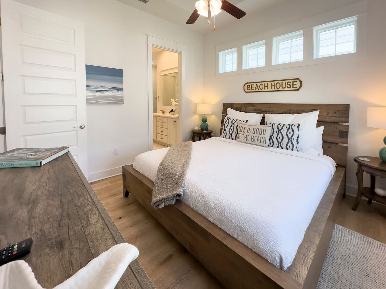 Une chambre avec un grand lit, un bureau en bois, un ventilateur de plafond et une vue sur la plage par la fenêtre.