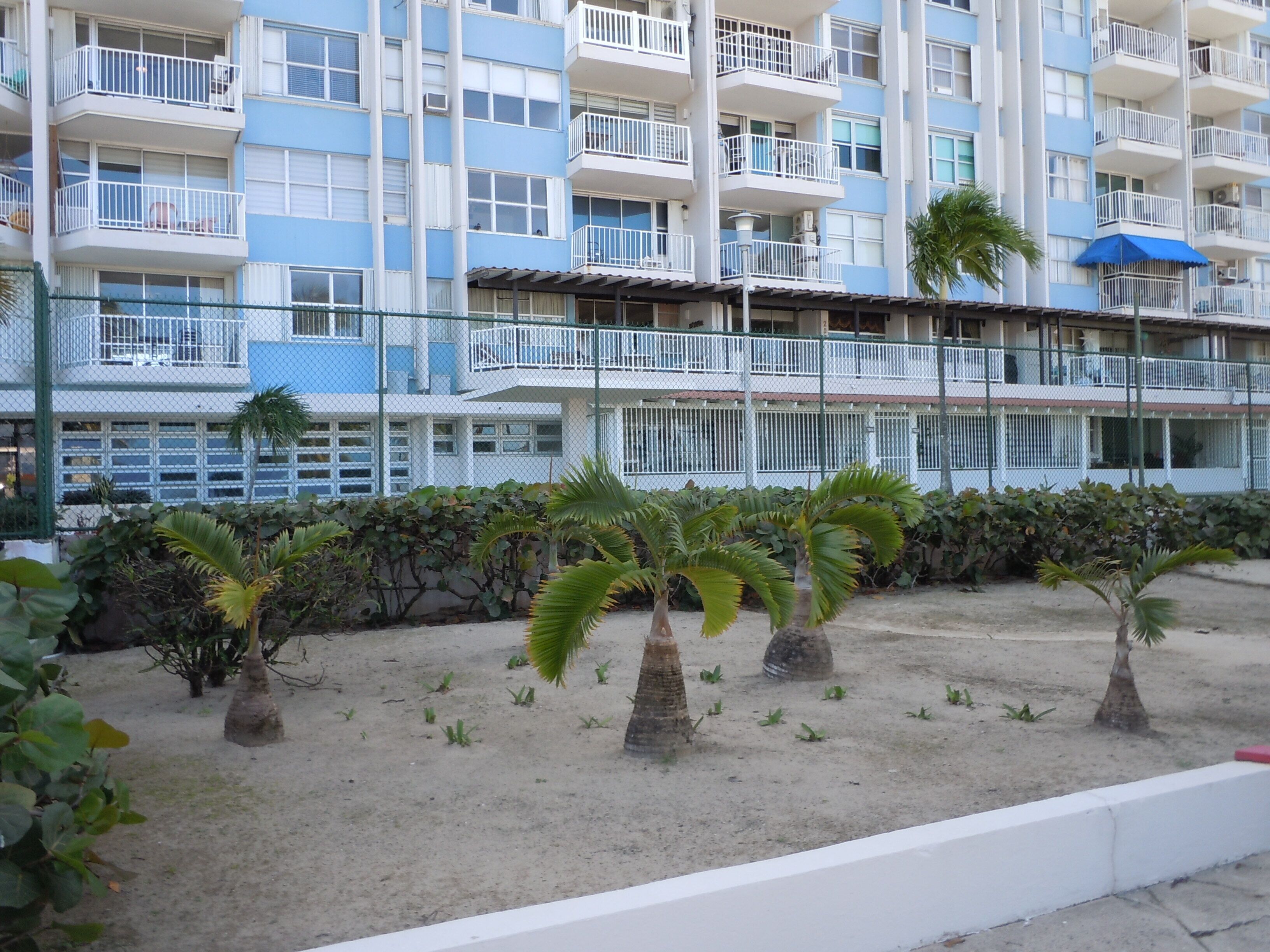 A multi-story building with balconies, a fenced garden area with palm trees, and a blue umbrella.