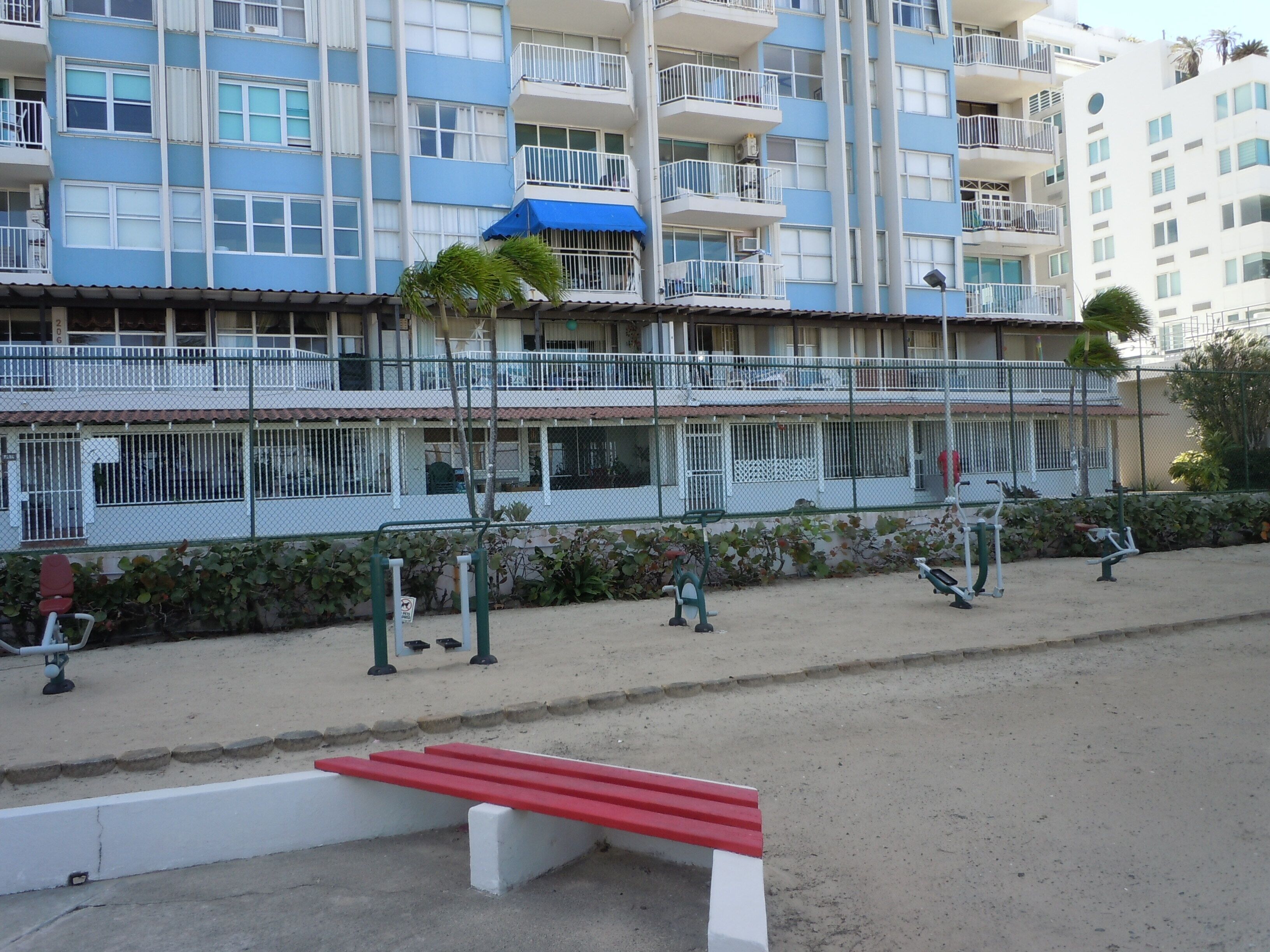 A multi-story residential building with balconies, a fenced outdoor area with exercise equipment, and a red bench.