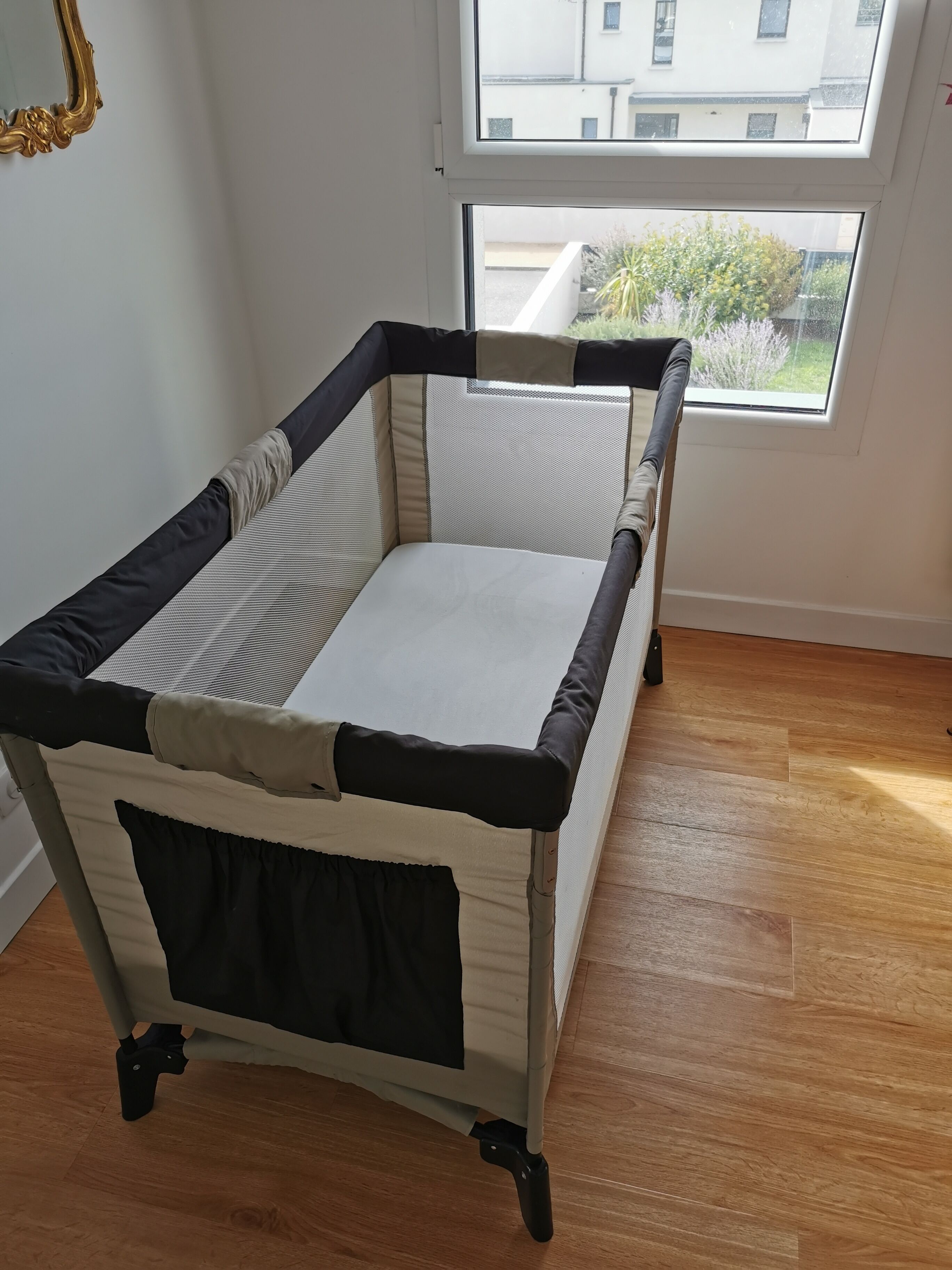 A foldable baby playpen with a mesh top and a cushioned bottom, positioned near a window with a view of buildings and greenery outside.