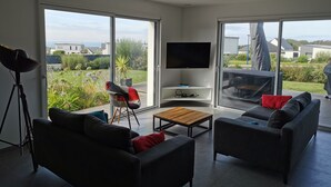 Een moderne woonkamer met een grote televisie, comfortabele zitplaatsen en uitzicht op een tuin en gebouwen.