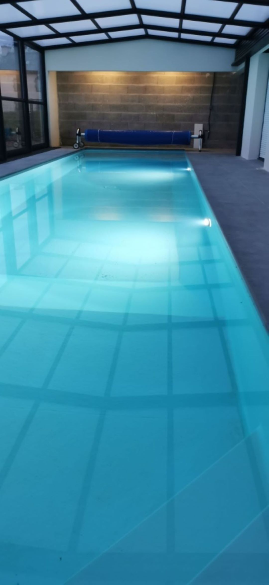 An indoor swimming pool with clear water, a tiled floor, and a brick wall in the background.