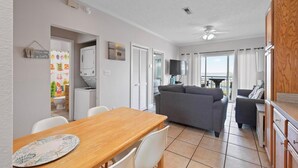 A modern living room with a grey sofa, a wooden dining table with white chairs, and a kitchen area with a TV and cabinets. - Quaint Beach Condo with Bay Views, Covered Parking, Pool, Bay Beach -Baywatch C4 (Pensacola Beach)