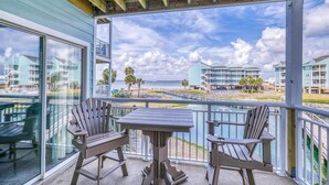 A balcony with Adirondack chairs and a table, overlooking a beach and buildings. - Quaint Beach Condo with Bay Views, Covered Parking, Pool, Bay Beach -Baywatch C4 (Pensacola Beach)
