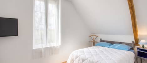 A bedroom with a sloped ceiling, a bed with white bedding, a bedside table with a lamp, and a window with sheer curtains.