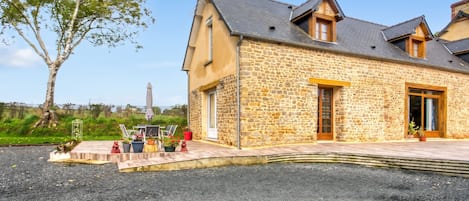 A stone building with a sloped roof, a paved driveway, and outdoor seating with chairs and umbrellas.