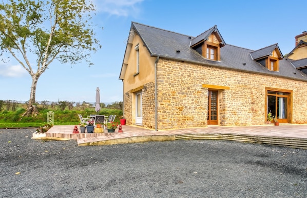 A stone building with a sloped roof, a paved driveway, and outdoor seating with chairs and umbrellas. - Homerez - House Saint-Marcouf (Saint-Marcouf)