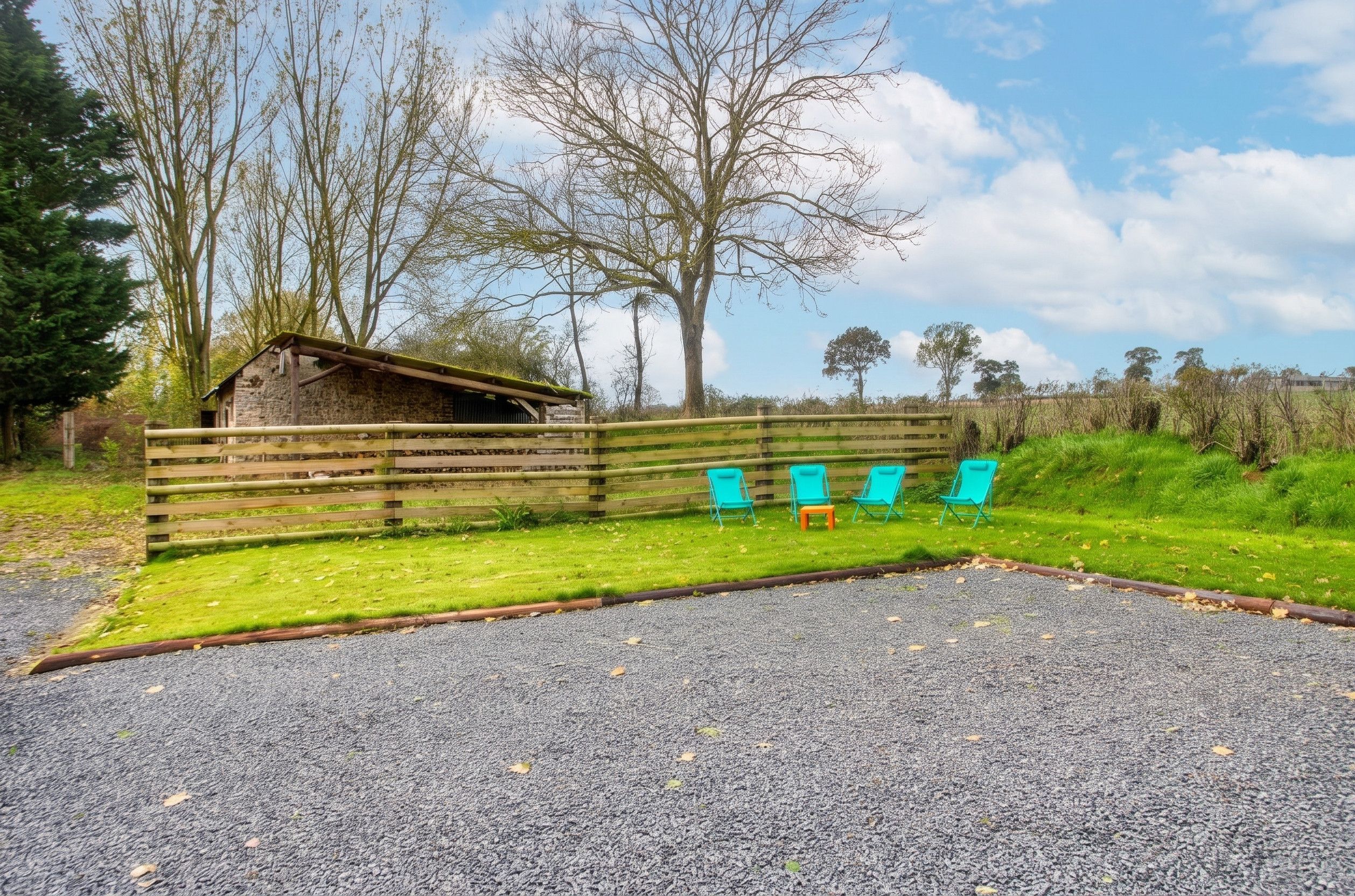 A gravel parking area with a wooden fence, a small stone building, and several blue chairs.