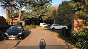 Une rue résidentielle bordée de voitures garées, avec une maison en briques et un grand arbre.