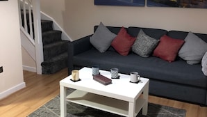Un salon avec un canapé gris foncé, une table basse blanche et un tapis gris. Un escalier se trouve à gauche, et deux tableaux sont accrochés au mur.