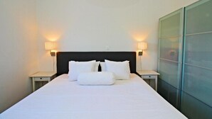 A neatly made bed with white linens and pillows, two bedside lamps, and a glass partition wall. - Luxury Apartments Gold and Grey (Zadar)