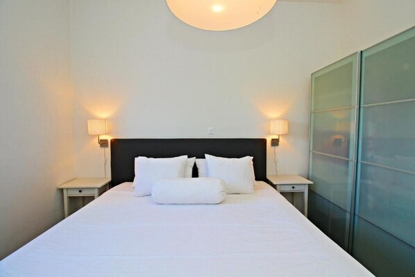 A neatly made bed with white linens and pillows, two bedside lamps, and a glass partition wall. - Luxury Apartments Gold and Grey (Zadar)
