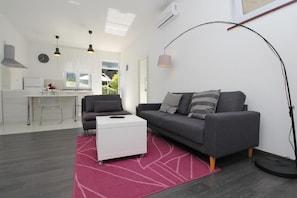 A modern living room with a grey sofa, a white coffee table, and a pink rug. There is a kitchen area with a white countertop and a chair.