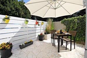 An outdoor patio with a table and chairs under a large umbrella, surrounded by potted plants and a white fence.