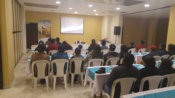 Uma sala de conferências com fileiras de cadeiras e mesas de plástico branco, uma tela de projetor e um microfone em um suporte.