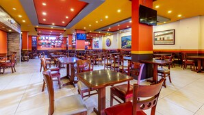A spacious restaurant with multiple tables and chairs, a TV mounted on the wall, and a colorful mural on the back wall. - Hotel Basan Plaza (San Juan de Pasto)