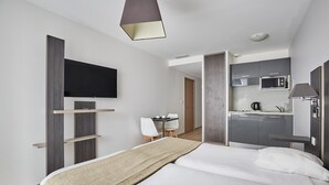 A modern hotel room with a bed, a TV mounted on the wall, a kitchenette, and a separate seating area with a table and chairs.