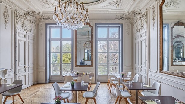 A dining room with ornate white walls, large windows, and a chandelier. Tables are set with chairs and centerpieces.