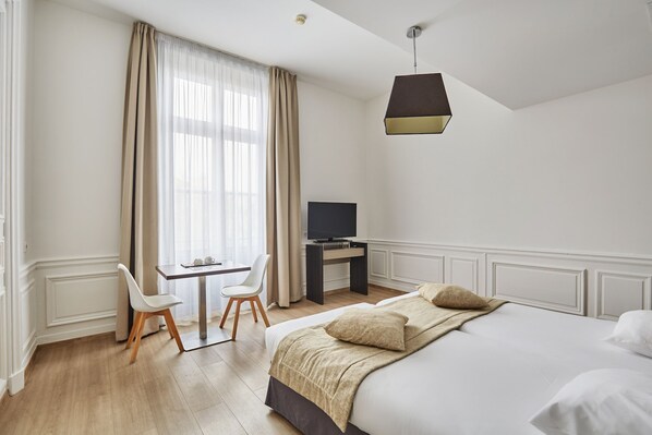 A hotel room with a bed, a desk with a TV, a chair, and a window with curtains.