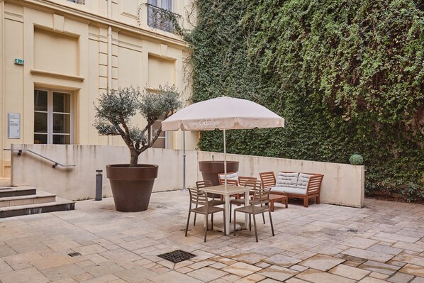 An outdoor seating area with a table and chairs, a large potted tree, and a wall covered in climbing plants.