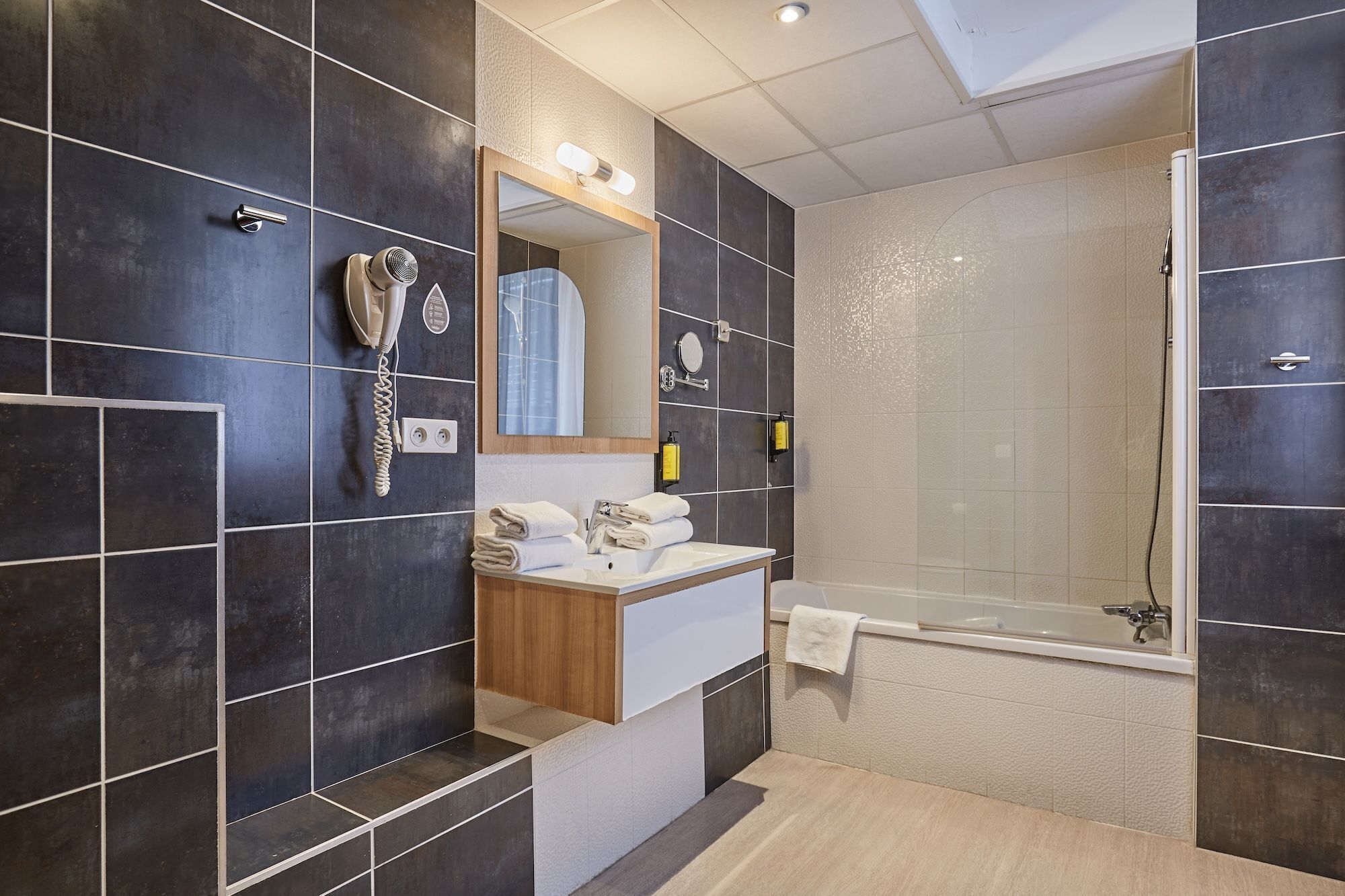 Une salle de bain moderne avec des carreaux foncés, un lavabo intégré, un miroir, un sèche-cheveux et une douche.