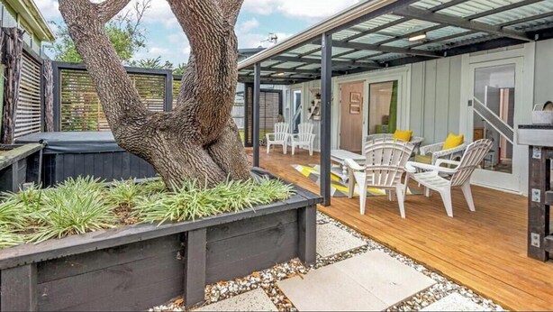 A covered outdoor area with a wooden deck, white chairs, and a large tree planter with greenery.