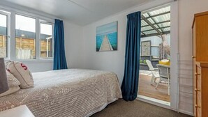 A bedroom with a bed, a desk, a chair, a window, a painting, and a door leading to an outdoor area.