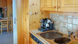 A kitchen with wooden cabinets, a coffee machine, a sink, and a dining area with chairs and a table. - Montagnards 17 by Interhome (Villars)