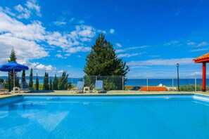 A swimming pool with lounge chairs and umbrellas, surrounded by a fence and trees, with a view of the ocean and mountains in the background.
