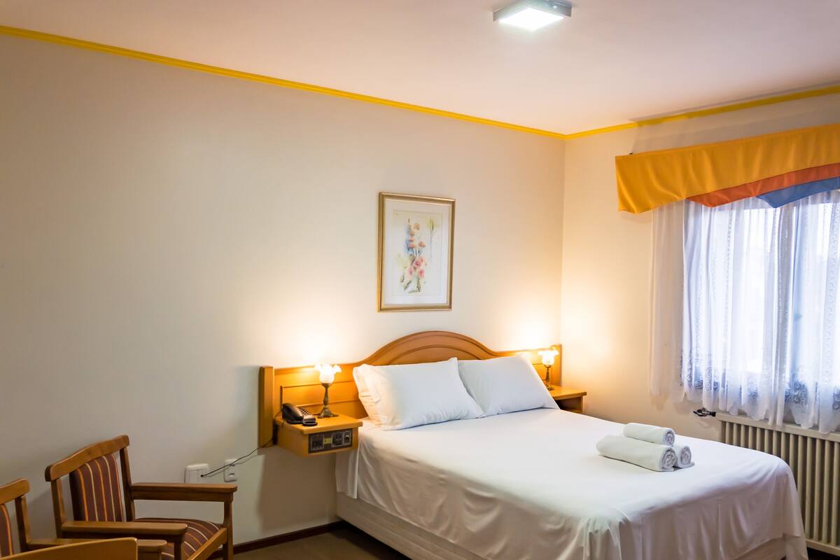 Habitación de hotel con cama, lámparas de noche, mesita de noche, una silla y una ventana con cortinas.