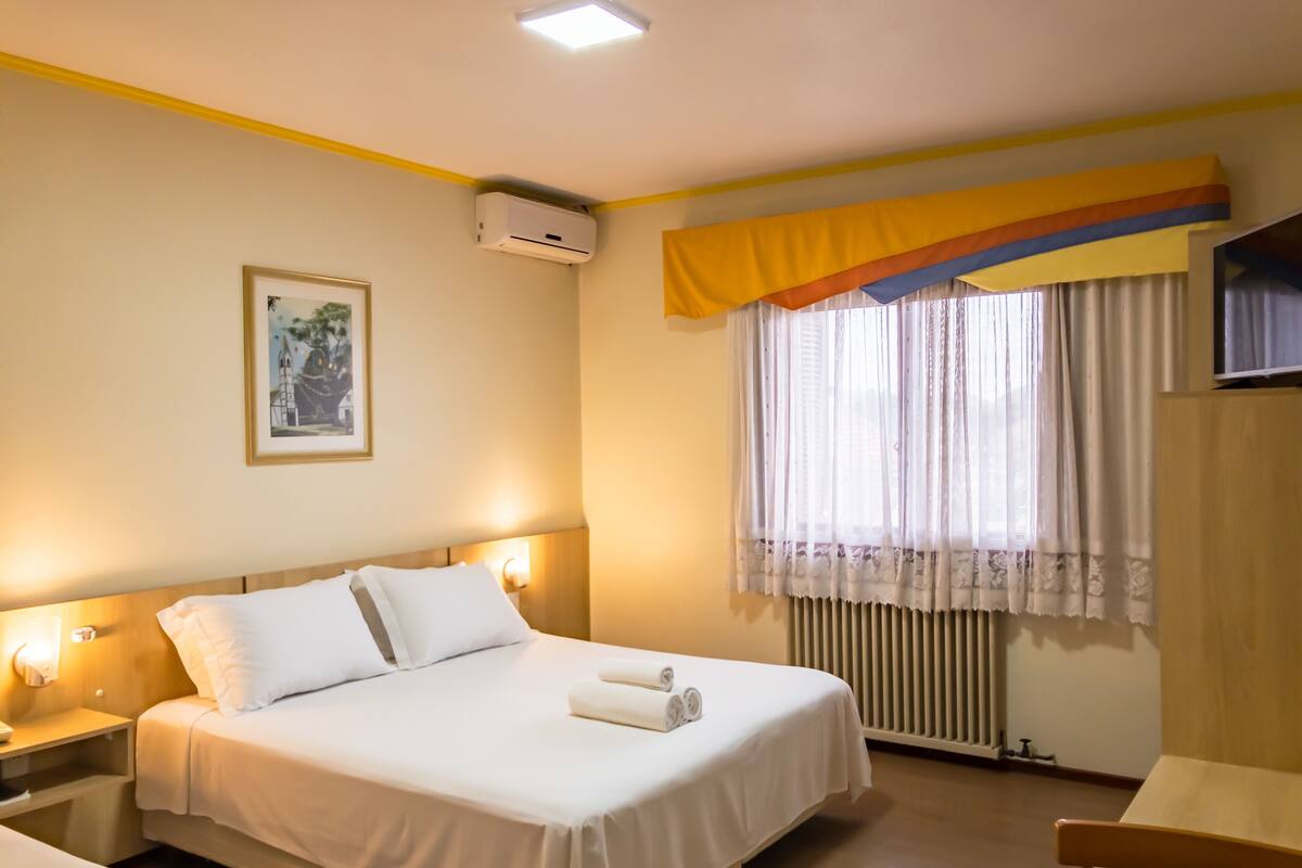 Una habitación de hotel con cama, lámparas de noche, ventana con cortinas, radiador y aire acondicionado instalado en la pared.