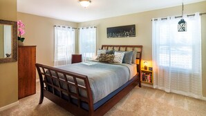 A bedroom with a wooden bed frame, a bedside table with a lamp, and a window with sheer curtains.