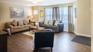 A living room with a beige sofa, a dark sofa, a coffee table, and a black chair. There is a lamp on a side table, a framed picture on the wall, and a window with curtains.