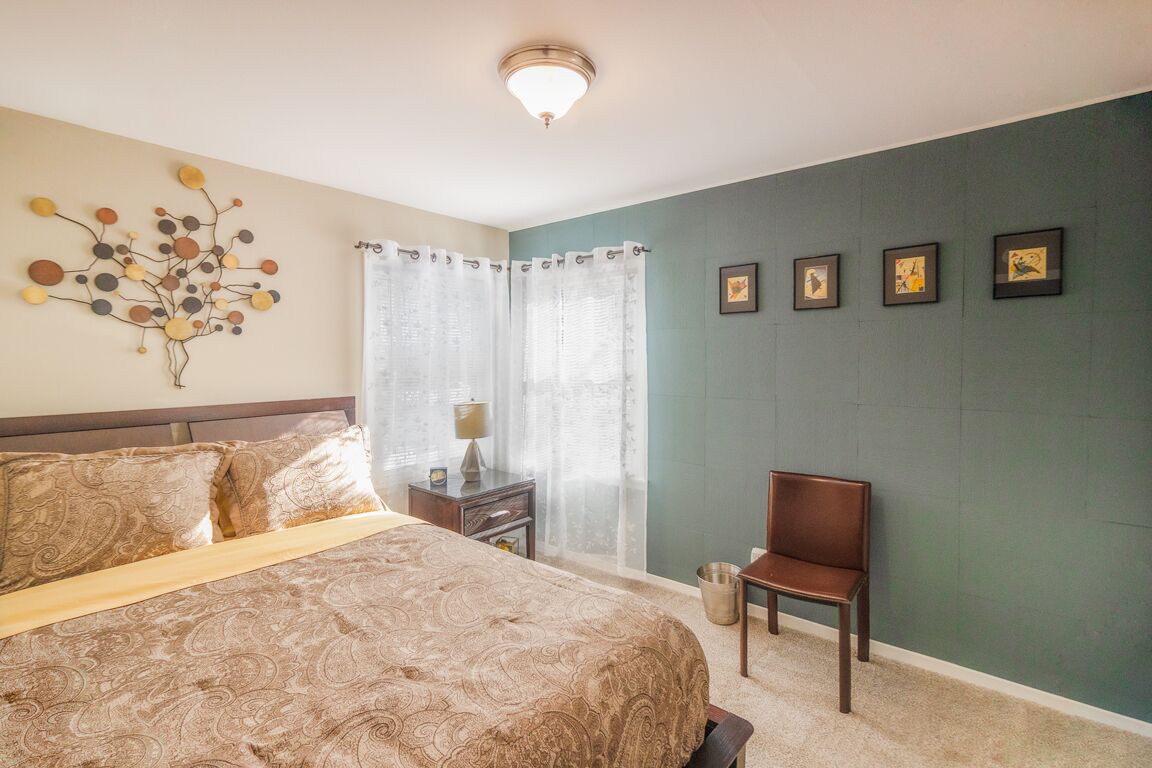 Un dormitorio con una cama grande, una silla, una mesita de noche, una lámpara y una pared con cuadros enmarcados.