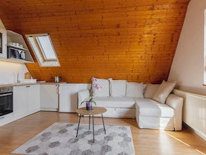 A cozy loft bedroom with a wooden ceiling, a white sofa, a small round table, and a kitchen area with white cabinets and appliances.