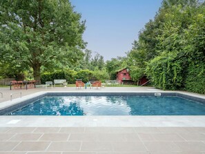 Une piscine rectangulaire, bordée d’une terrasse carrelée, entourée d’arbres et d’un bâtiment rouge.