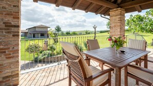 A wooden deck with a table and chairs, overlooking a green landscape with a house in the background. - Beautiful  villa for 4 people with WIFI, private pool, TV, terrace and panoramic view (Castagnole Delle Lanze)