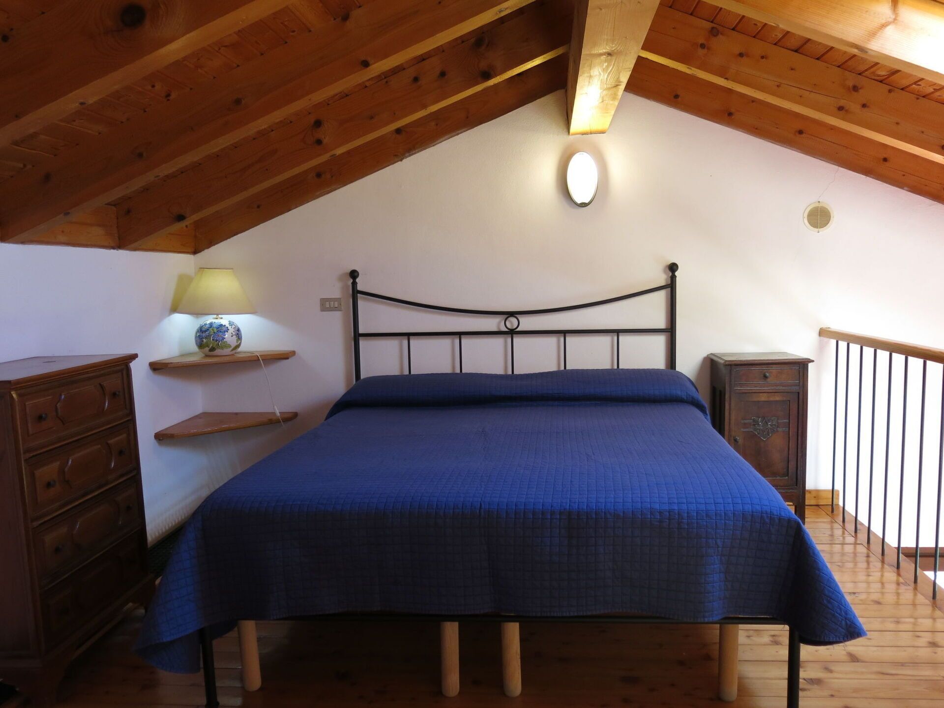 A bedroom with a sloped ceiling, a metal bed frame, a blue bedspread, wooden furniture, and a small bedside table with a lamp.