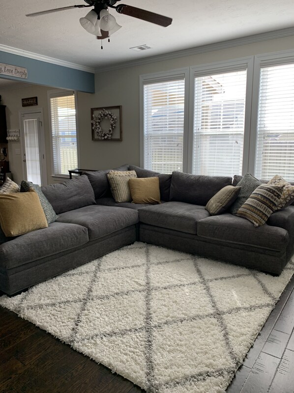 A living room with a gray sectional sofa, a fluffy white rug, and a ceiling fan.