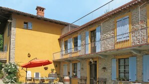 Un patio con una mesa y sillas, un paraguas rojo y un edificio de piedra con contraventanas azules.