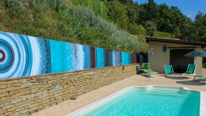 Una piscina con un muro de piedra y un mural. Hay sillones de descanso y un área techada con una sombrilla.