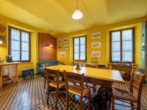 Uma sala de jantar com uma mesa comprida, cadeiras de madeira e um banco. Há janelas com cortinas, um lustre e obras de arte penduradas na parede.