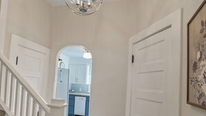 A hallway with a staircase, a chandelier, and a door leading to a kitchen area. - The Comfy Craftsman l KING Beds l Near Park l Spacious Home (Spokane)